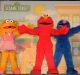 Three Sesame Street characters stand together with their arms open wide. From left to right, they are a yellow character wearing a pink dress, a red character, and a blue character. The backdrop resembles a colorful Sesame Street neighborhood, showcasing the charm of kids' programming by Sesame Workshop.