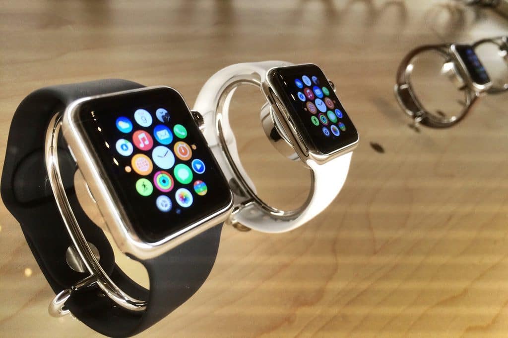 Three smartwatches, including an Apple Watch Series 3 with Cellular, are displayed on a wooden surface. The two closest watches have black and white bands, respectively, and their screens show various colorful app icons. The third watch is further in the background and slightly out of focus.