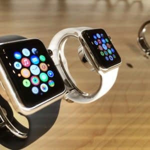 Three smartwatches, including an Apple Watch Series 3 with Cellular, are displayed on a wooden surface. The two closest watches have black and white bands, respectively, and their screens show various colorful app icons. The third watch is further in the background and slightly out of focus.