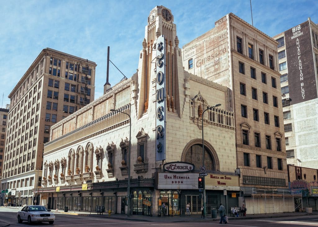 A historic theatre building stands at a street corner in an urban area, now housing a bustling retail store. The tall, ornate facade features intricate architectural details and a vertical sign with the word "Tower." Nearby, pedestrians and a few cars are visible against a backdrop of adjacent buildings.