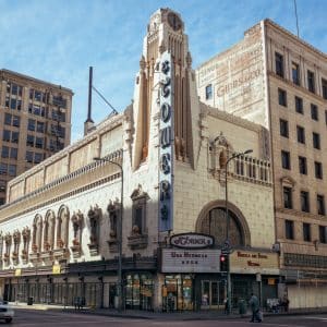 A historic theatre building stands at a street corner in an urban area, now housing a bustling retail store. The tall, ornate facade features intricate architectural details and a vertical sign with the word "Tower." Nearby, pedestrians and a few cars are visible against a backdrop of adjacent buildings.