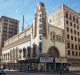 A historic theatre building stands at a street corner in an urban area, now housing a bustling retail store. The tall, ornate facade features intricate architectural details and a vertical sign with the word "Tower." Nearby, pedestrians and a few cars are visible against a backdrop of adjacent buildings.