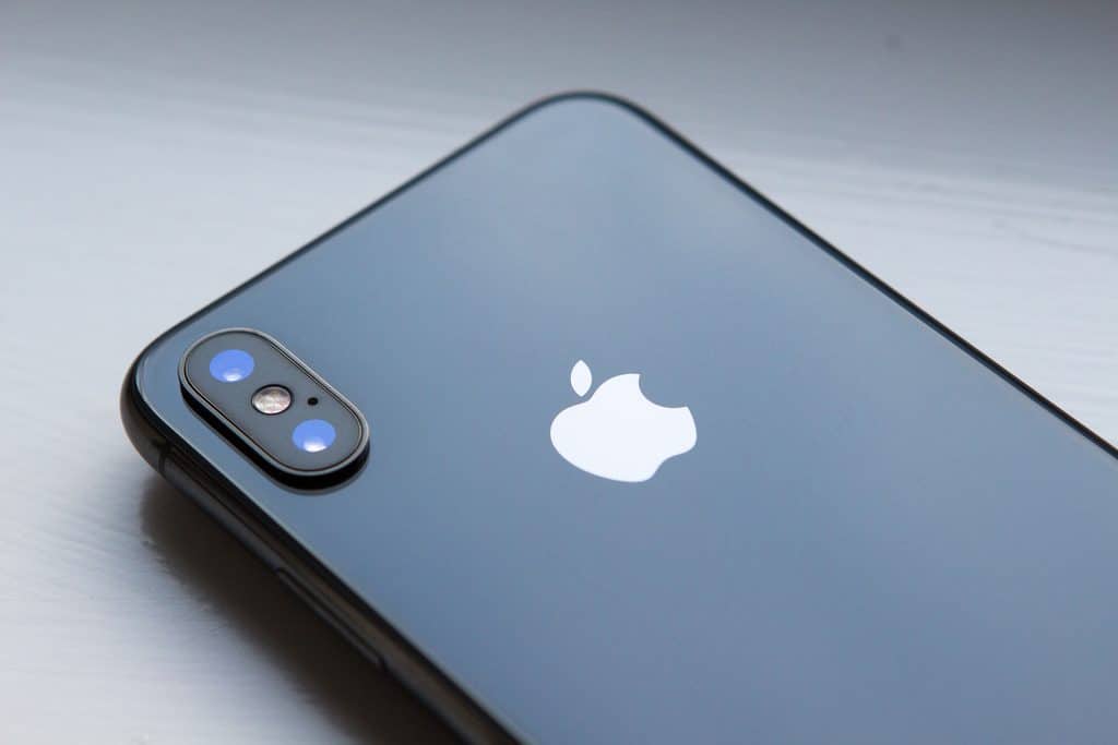 A close-up of the back of a gray iPhone with a shiny surface. The Apple logo is prominently displayed in the center. The dual camera setup with flash is visible in the top left corner of the phone, highlighting its advanced security features powered by iOS 12. The phone is placed on a white, smooth surface.