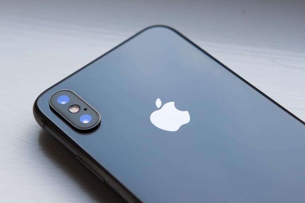 A close-up of the back of a gray iPhone with a shiny surface. The Apple logo is prominently displayed in the center. The dual camera setup with flash is visible in the top left corner of the phone, highlighting its advanced security features powered by iOS 12. The phone is placed on a white, smooth surface.