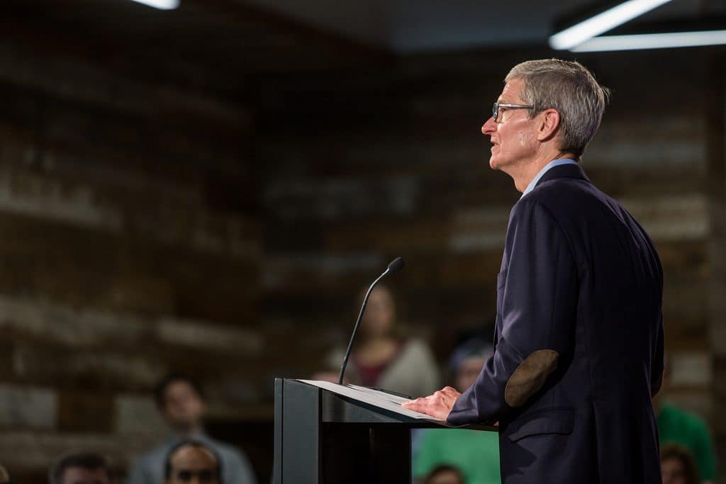 A man in a business suit stands at a podium, speaking into a microphone. He is addressing the audience on matters of privacy, which appears out of focus in the background. The setting looks like an indoor event or conference, with wooden paneling on the walls.