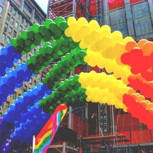 A colorful arch made of balloons in rainbow colors is displayed outdoors in an urban setting. Buildings and construction scaffolding are visible in the background, along with a smaller rainbow flag near the balloon arch, celebrating Pride month with vibrant inclusion perfect for social media updates.