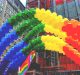 A colorful arch made of balloons in rainbow colors is displayed outdoors in an urban setting. Buildings and construction scaffolding are visible in the background, along with a smaller rainbow flag near the balloon arch, celebrating Pride month with vibrant inclusion perfect for social media updates.