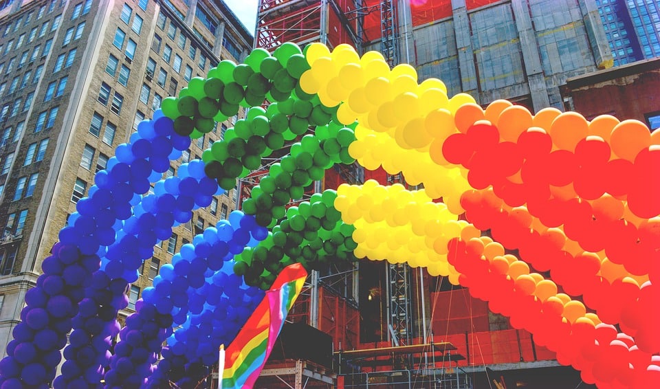 A colorful arch made of balloons in rainbow colors is displayed outdoors in an urban setting. Buildings and construction scaffolding are visible in the background, along with a smaller rainbow flag near the balloon arch, celebrating Pride month with vibrant inclusion perfect for social media updates.