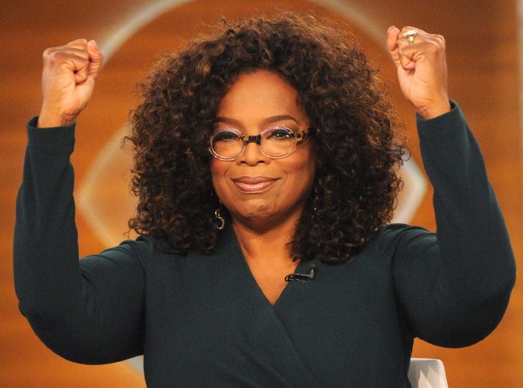 A person with curly hair, wearing glasses and a dark green top, raises both fists in the air with a confident smile. The background is a warm, orange-toned setting that gives off major Oprah Winfrey vibes.