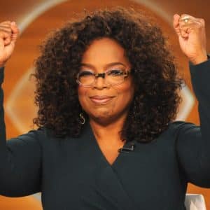 A person with curly hair, wearing glasses and a dark green top, raises both fists in the air with a confident smile. The background is a warm, orange-toned setting that gives off major Oprah Winfrey vibes.