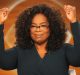 A person with curly hair, wearing glasses and a dark green top, raises both fists in the air with a confident smile. The background is a warm, orange-toned setting that gives off major Oprah Winfrey vibes.