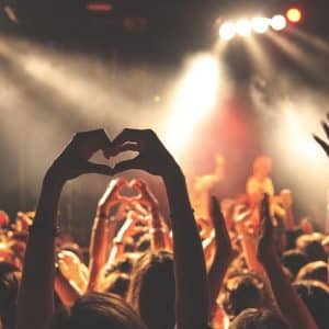 A crowd of people at the BottleRock music festival, with hands raised and some forming heart shapes. Bright stage lights illuminate the scene, casting a vibrant atmosphere. The background is slightly blurred, focusing on the energetic audience in the foreground.