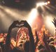 A crowd of people at the BottleRock music festival, with hands raised and some forming heart shapes. Bright stage lights illuminate the scene, casting a vibrant atmosphere. The background is slightly blurred, focusing on the energetic audience in the foreground.