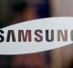 A close-up image of the Samsung logo on a transparent surface. The word "SAMSUNG" is written in bold, black capital letters inside a white, elongated oval shape, displayed against a blurred background that hints at the anticipating launch of the Samsung Galaxy Note 9.