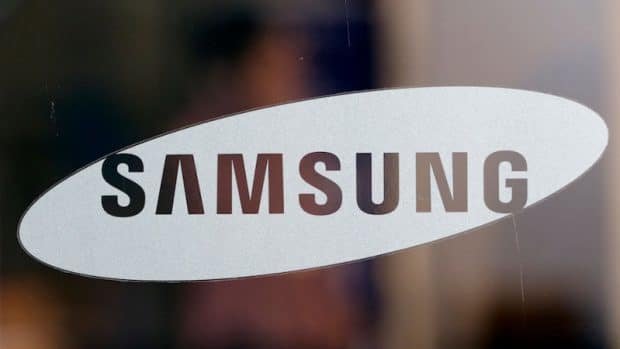 A close-up image of the Samsung logo on a transparent surface. The word "SAMSUNG" is written in bold, black capital letters inside a white, elongated oval shape, displayed against a blurred background that hints at the anticipating launch of the Samsung Galaxy Note 9.