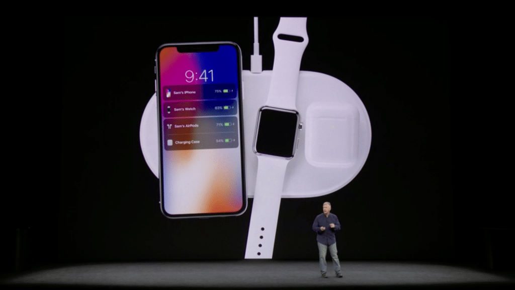 A person stands on stage presenting Apple's latest release, a charging pad that simultaneously powers a smartphone, smartwatch, and wireless earbuds. The smartphone screen displays it is charging at 78%, the smartwatch at 61%, and the earbuds case at 48%. This innovation recalls the ambition of AirPower.