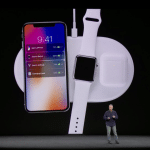 A person stands on stage presenting Apple's latest release, a charging pad that simultaneously powers a smartphone, smartwatch, and wireless earbuds. The smartphone screen displays it is charging at 78%, the smartwatch at 61%, and the earbuds case at 48%. This innovation recalls the ambition of AirPower.