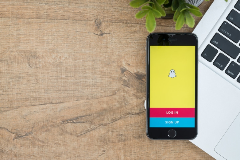 A smartphone displaying a yellow screen with a ghost icon, signaling the launch of Snapchat, offers "Log In" and "Sign Up" options. It's placed on a wooden desk next to a silver laptop, while a small green potted plant is partially visible at the top right corner.