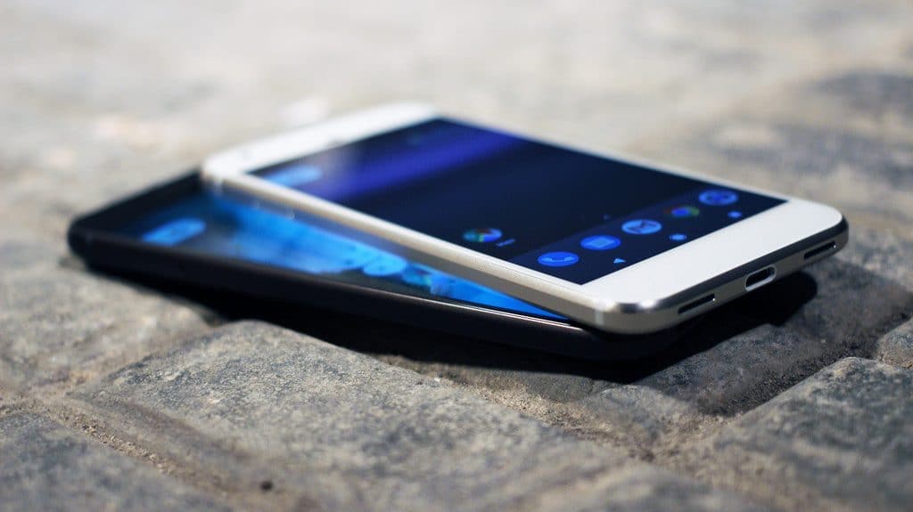 Two smartphones are placed on top of each other on a textured gray surface. The front phone, potentially a Pixel 3 from 2018, showcases app icons, including a blue and green icon and a green and white icon. The phones are angled, with the front phone appearing slightly raised, exemplifying its sleek design.