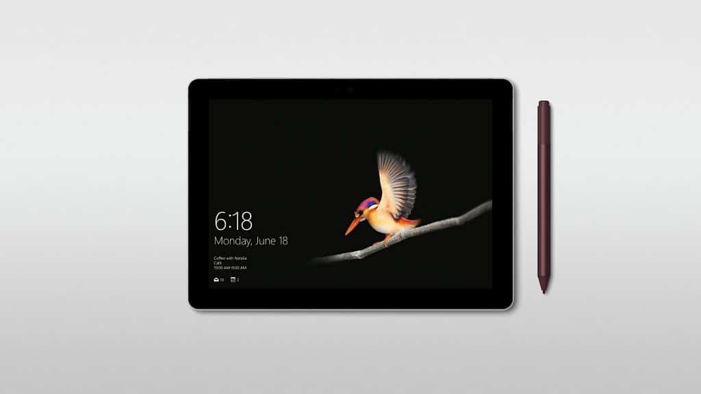 A Microsoft Surface Go on a white background, displaying the lock screen. The screen shows a bird mid-flight against a black background with the time of 6:18 and the date Monday, June 18. A stylus is placed vertically next to the tablet on the right.