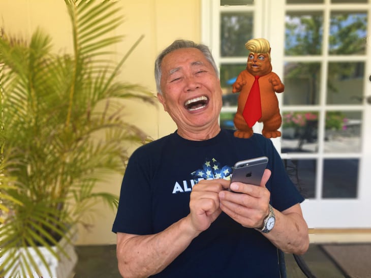 A man with gray hair and glasses is laughing while looking at his smartphone. Behind him is an animated character resembling a small, caricatured figure with blonde hair, wearing a red tie and suit, standing near a potted plant and a glass door. The scene has an amusingly surreal quality, almost like he's discovered the latest Trumpy Cat meme.