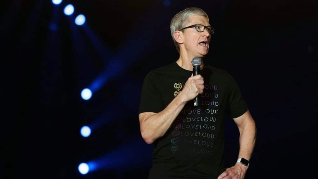 A person with short gray hair and glasses is speaking into a microphone while standing on a stage. They are wearing a black t-shirt with the word "LOVELOUD" repeated in various colors, delivering a message of hope to LGBTQ+ youth. The background is dark with several lit spotlights visible.