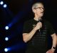 A person with short gray hair and glasses is speaking into a microphone while standing on a stage. They are wearing a black t-shirt with the word "LOVELOUD" repeated in various colors, delivering a message of hope to LGBTQ+ youth. The background is dark with several lit spotlights visible.