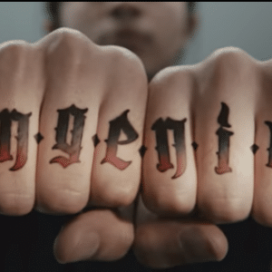 A close-up image of a person's knuckles with tattoos that spell out the word "ingenius" in a gothic font. The hand is clenched into fists, with the background blurred to emphasize the tattooed letters, reminiscent of a rebellious Samsung ad.