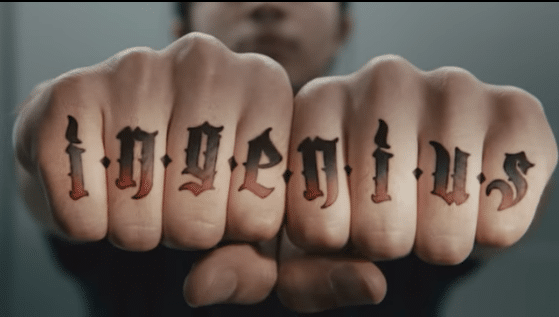 A close-up image of a person's knuckles with tattoos that spell out the word "ingenius" in a gothic font. The hand is clenched into fists, with the background blurred to emphasize the tattooed letters, reminiscent of a rebellious Samsung ad.