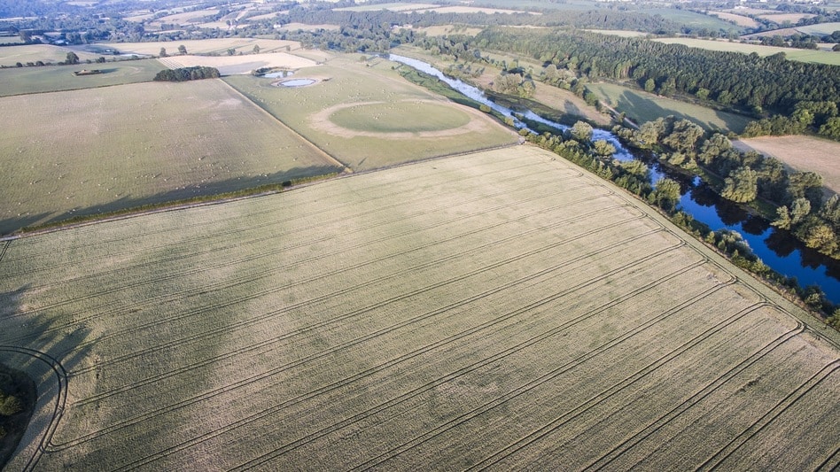 Aerial view of a vast rural landscape featuring large agricultural fields, a meandering river, and patches of woodland. The scene shows a mix of green pastures and cultivated land, with the river winding through the countryside. Visible amid Europe's heatwave is a mysterious henge emerging from parched earth.