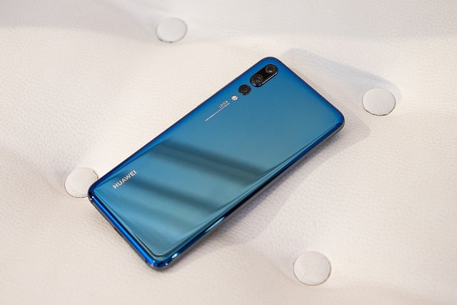 A blue Huawei smartphone, highlighted amid rising smartphone sales, is shown lying face down on a white, quilted surface. The back of the phone features the brand name "Huawei" and a triple-lens camera setup with "Leica" branding.