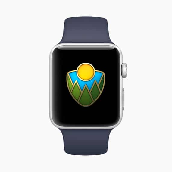 A smartwatch with a dark blue band displays a hiking badge on its screen. The badge showcases a stylized sun above green mountains against a blue sky on a black background, celebrating National Parks. This device also supports Apple Pay for added convenience on your adventures.