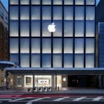 A stunning modern storefront with a prominent illuminated Apple logo at the top, large glass windows displaying the interior of the Kyoto Apple store. The exterior is sleek and minimalistic, with a pedestrian walkway in front and a street with blurred car lights indicating motion.