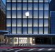 A stunning modern storefront with a prominent illuminated Apple logo at the top, large glass windows displaying the interior of the Kyoto Apple store. The exterior is sleek and minimalistic, with a pedestrian walkway in front and a street with blurred car lights indicating motion.