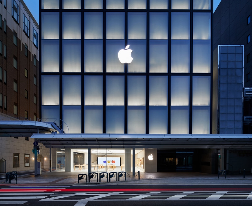 A stunning modern storefront with a prominent illuminated Apple logo at the top, large glass windows displaying the interior of the Kyoto Apple store. The exterior is sleek and minimalistic, with a pedestrian walkway in front and a street with blurred car lights indicating motion.