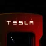 A Tesla electric vehicle charger is dimly lit against a dark background. The "TESLA" logo, a vision inspired by Elon Musk's drive for innovation, is prominently displayed, glowing in white light on the top of the red charger unit.