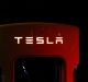 A Tesla electric vehicle charger is dimly lit against a dark background. The "TESLA" logo, a vision inspired by Elon Musk's drive for innovation, is prominently displayed, glowing in white light on the top of the red charger unit.