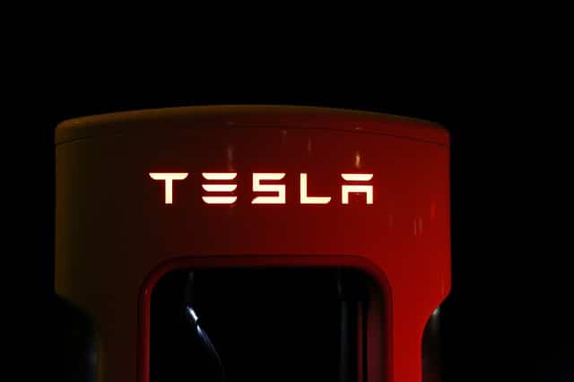 A Tesla electric vehicle charger is dimly lit against a dark background. The "TESLA" logo, a vision inspired by Elon Musk's drive for innovation, is prominently displayed, glowing in white light on the top of the red charger unit.