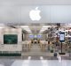 An expanded Australian Apple Store with a large Apple logo above it showcases an interior lit by white lights. The entrance features a display table with a MacBook Pro and various tech gadgets. The side window displays numerous app icons, while inside, rows of tables hold an array of devices.