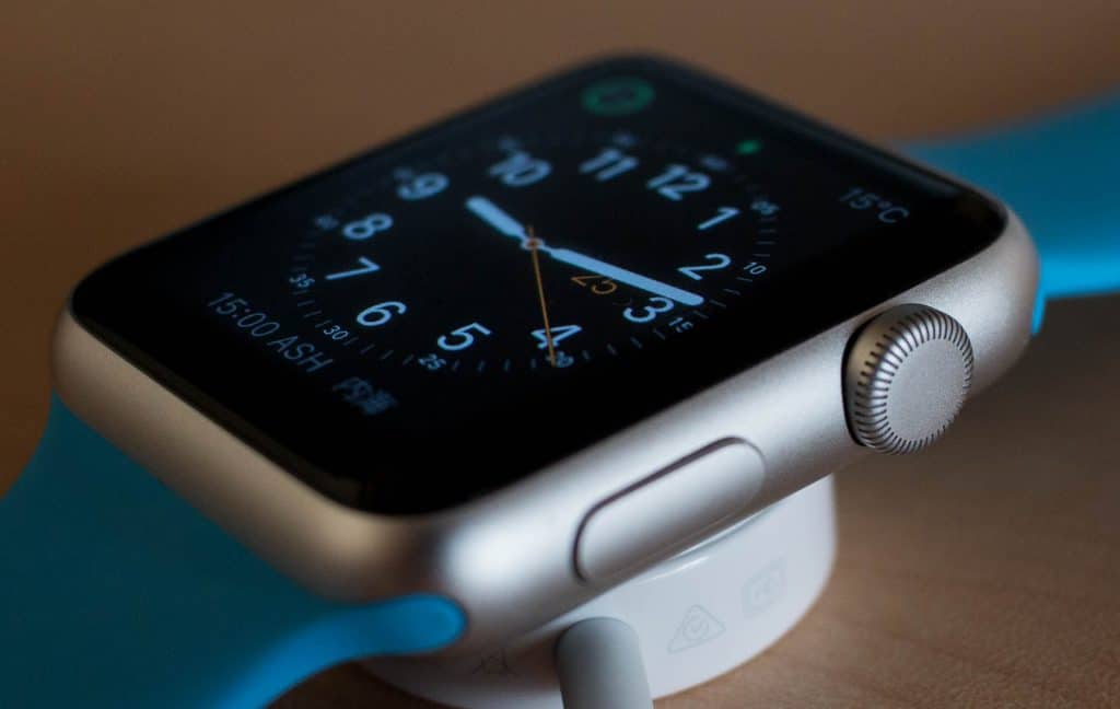 A close-up of a silver smartwatch with a blue strap, displaying an analog clock face on its screen. The watch, similar to popular Apple Watches everyone loves, is lying on its side on a light-colored surface, engaged in charging via a cable connected to a circular magnetic charger.