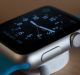 A close-up of a silver smartwatch with a blue strap, displaying an analog clock face on its screen. The watch, similar to popular Apple Watches everyone loves, is lying on its side on a light-colored surface, engaged in charging via a cable connected to a circular magnetic charger.