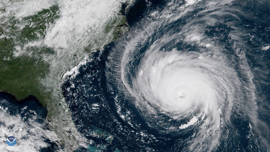 A satellite image shows Hurricane Florence, a powerful storm with a well-defined eye, approaching the southeastern coast of the United States. The large, spiraling storm covers a substantial portion of the Atlantic Ocean, with the green coastline of the U.S. visible on the left.