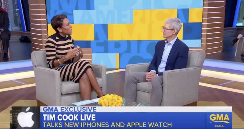 Two people sit in chairs facing each other on a TV set. The person on the left, Tim Cook, is gesturing with his hands while speaking, and the person on the right is listening attentively. The background displays "Good Morning America" and news graphics. A text reads "TIM COOK LIVE: Talks New iPhones and Apple Watch".