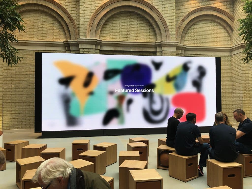 A group of people sits on wooden stools in front of a large screen displaying colorful abstract art and the text "Today at Apple Creative, Featured Sessions." The setting appears to be an indoor event space with brick walls and tall arches, likely a new Apple Store worth a visit.