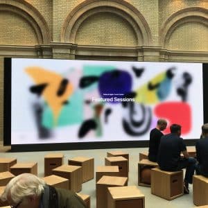 A group of people sits on wooden stools in front of a large screen displaying colorful abstract art and the text "Today at Apple Creative, Featured Sessions." The setting appears to be an indoor event space with brick walls and tall arches, likely a new Apple Store worth a visit.