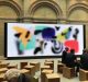 A group of people sits on wooden stools in front of a large screen displaying colorful abstract art and the text "Today at Apple Creative, Featured Sessions." The setting appears to be an indoor event space with brick walls and tall arches, likely a new Apple Store worth a visit.