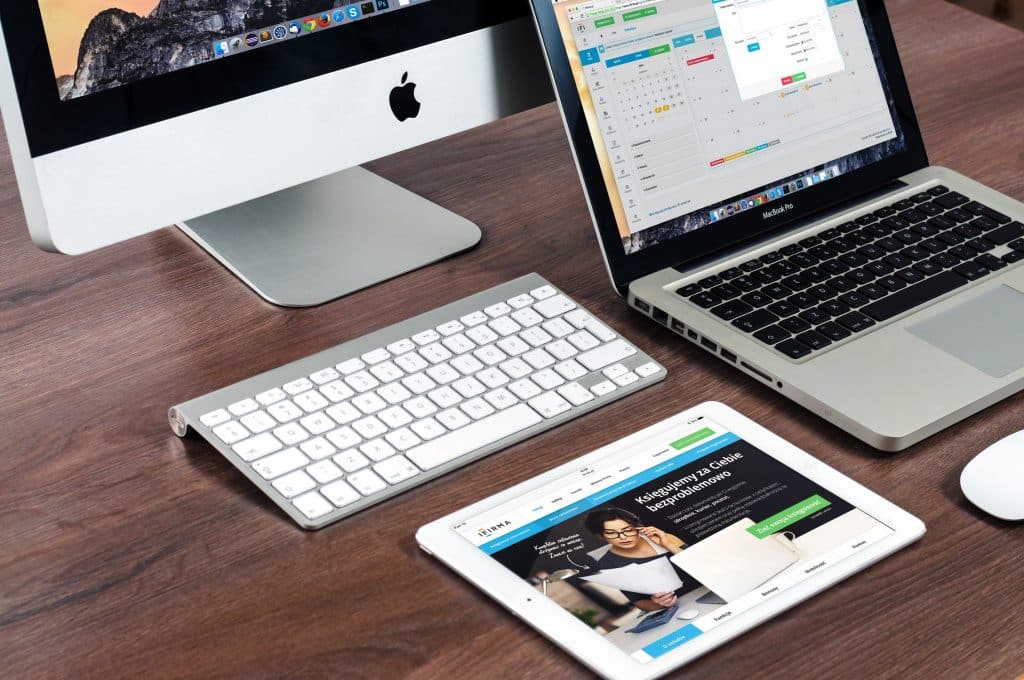 A wooden desk with an Apple desktop computer, a MacBook laptop, and an iPad. The desktop features a wireless keyboard and mouse. The laptop screen displays a calendar application, while the iPad shows a website homepage—hinting at new Mac models anticipated from the upcoming October event.