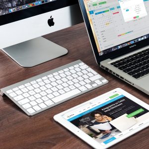 A wooden desk with an Apple desktop computer, a MacBook laptop, and an iPad. The desktop features a wireless keyboard and mouse. The laptop screen displays a calendar application, while the iPad shows a website homepage—hinting at new Mac models anticipated from the upcoming October event.