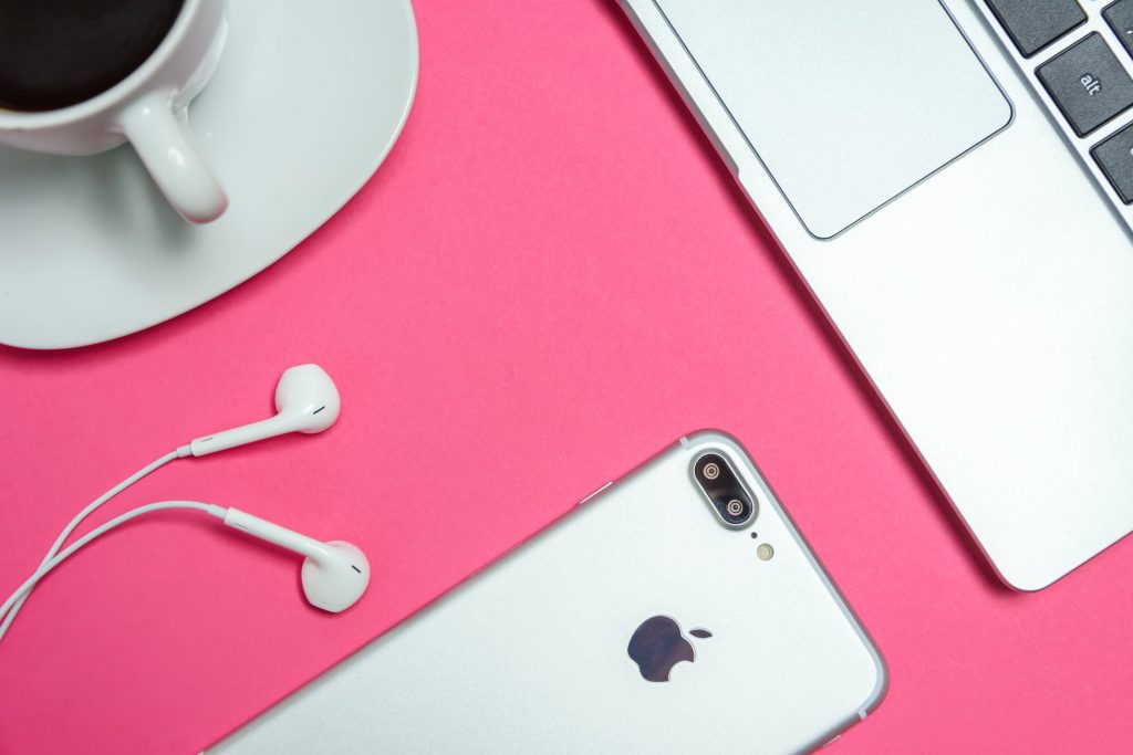 A white iPhone with earbuds, a white cup on a saucer, and a silver laptop are placed on a bright pink background. The setup evokes retro designs as the earbuds connect to the iPhone, and the cup is filled with dark coffee.