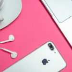 A white iPhone with earbuds, a white cup on a saucer, and a silver laptop are placed on a bright pink background. The setup evokes retro designs as the earbuds connect to the iPhone, and the cup is filled with dark coffee.